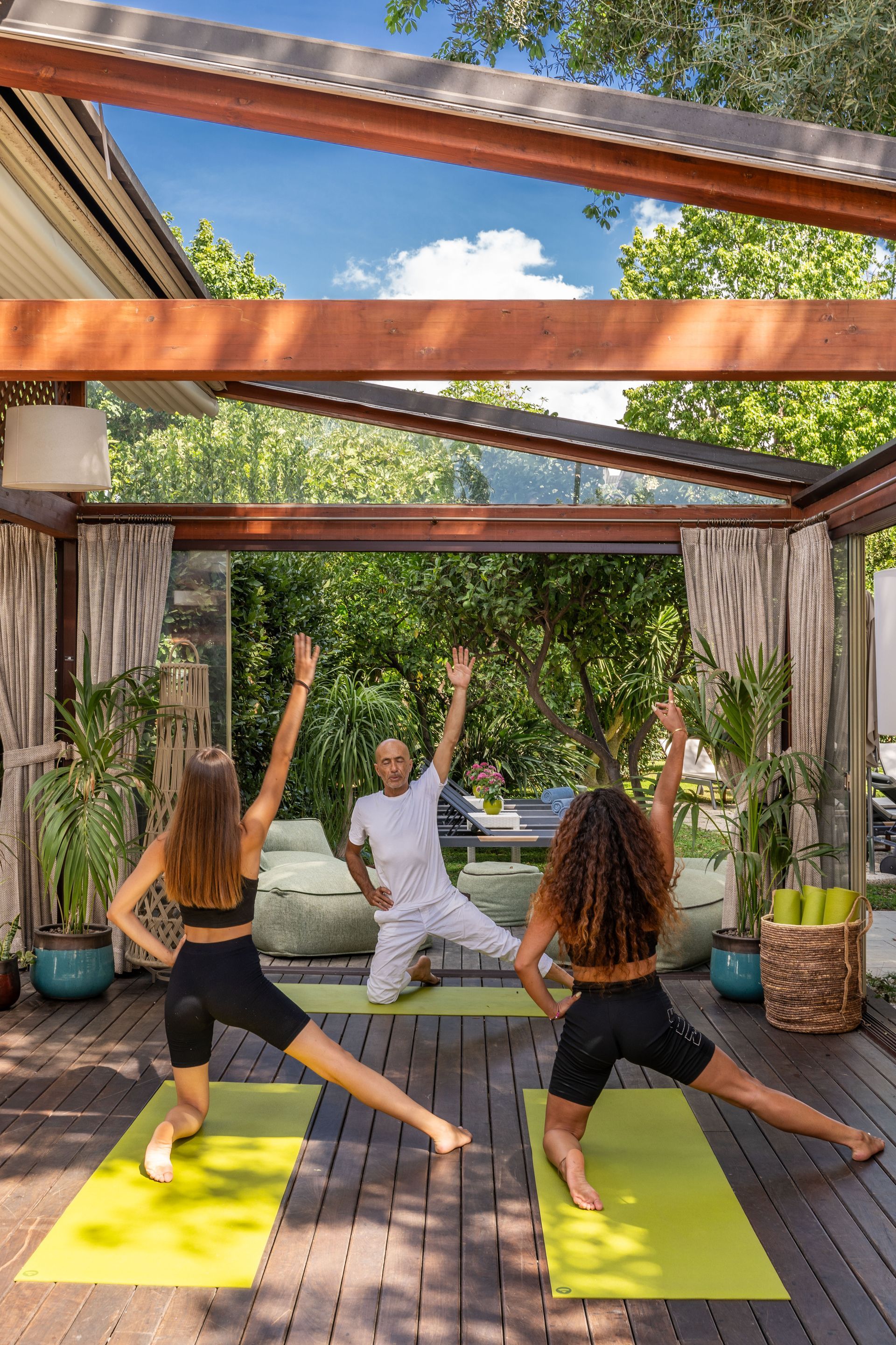 Three people perform a kneeling, arm-stretched yoga pose on green mats on a wooden deck in an outdoor garden setting.
