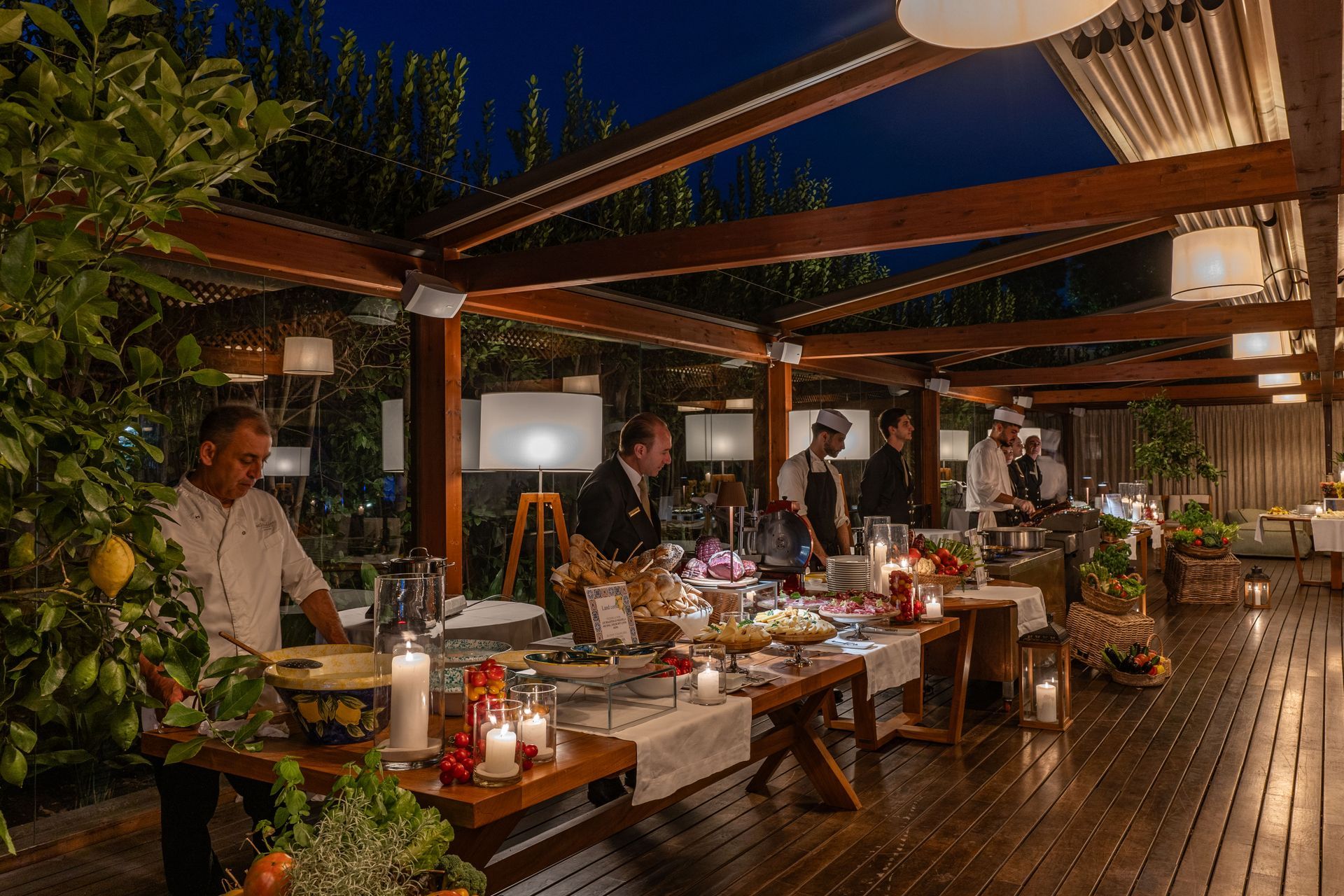 Chefs prepare a buffet on a candlelit wooden deck at night under a timber-framed patio structure with warm lighting.