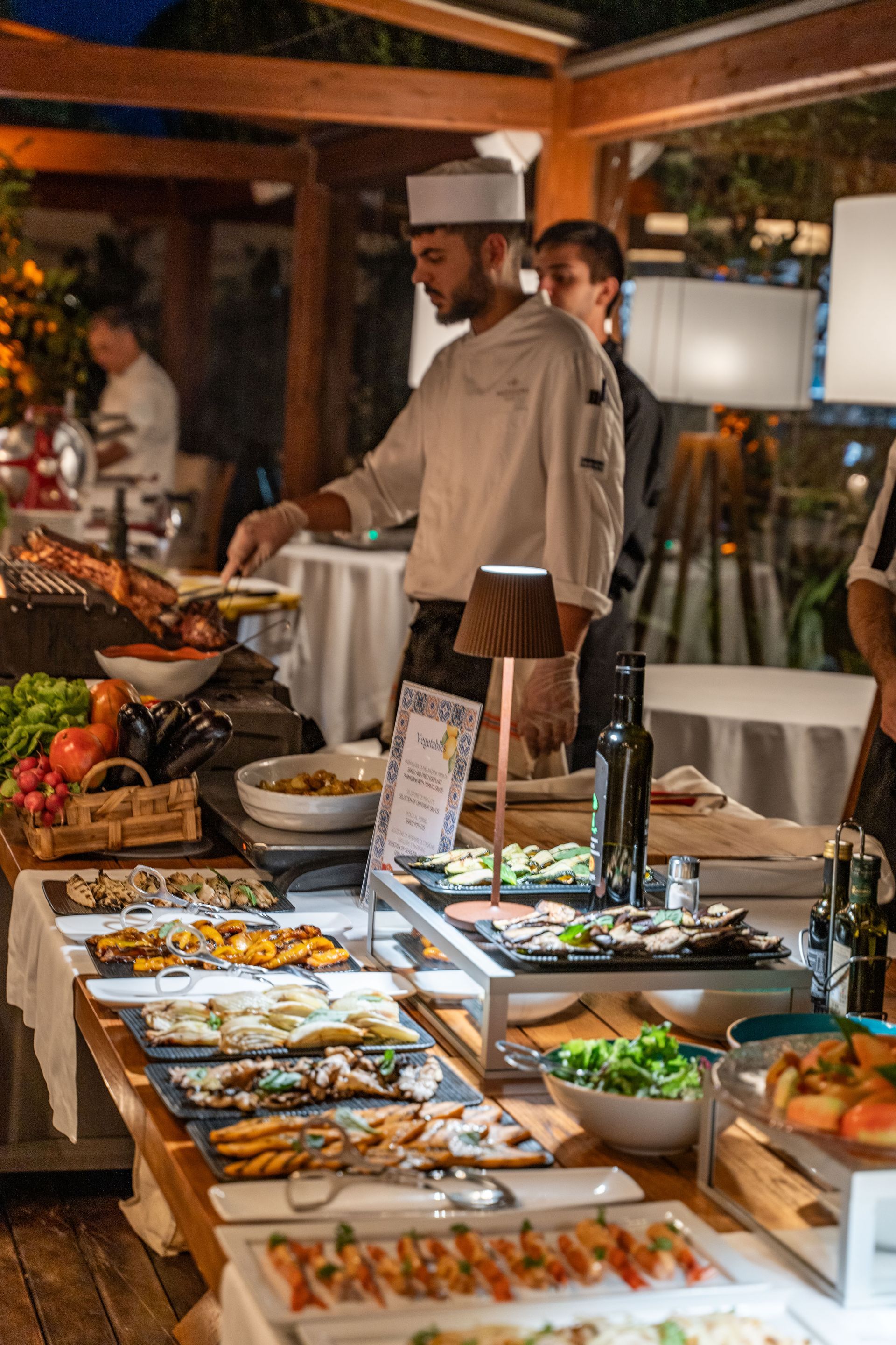 A chef in uniform prepares food at a restaurant buffet, with several platters of appetizers arranged on a long table.