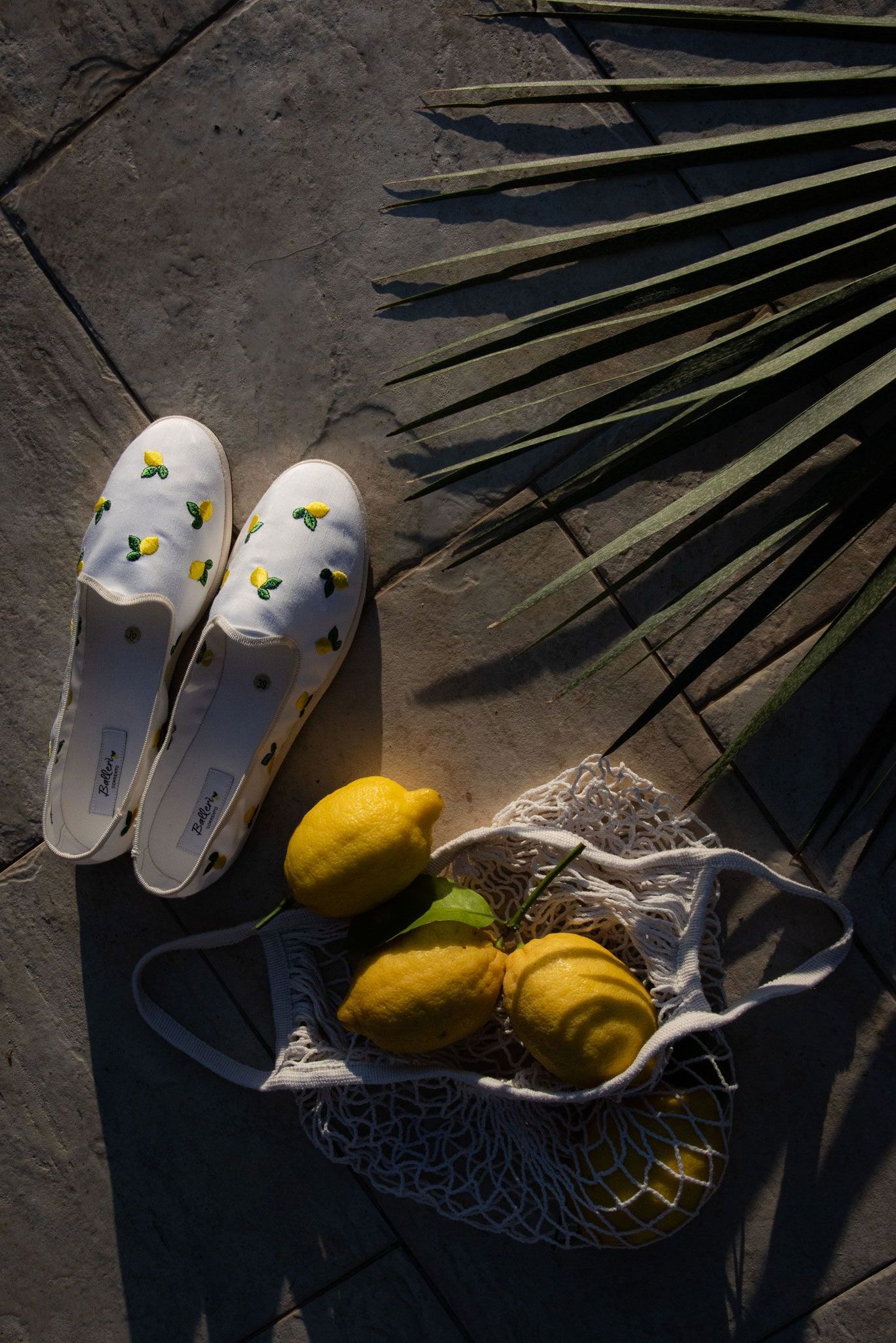 Espadrillas bianche con motivi di limoni, tre limoni gialli in una borsa all'uncinetto e una fronda di palma su una superficie di pietra.