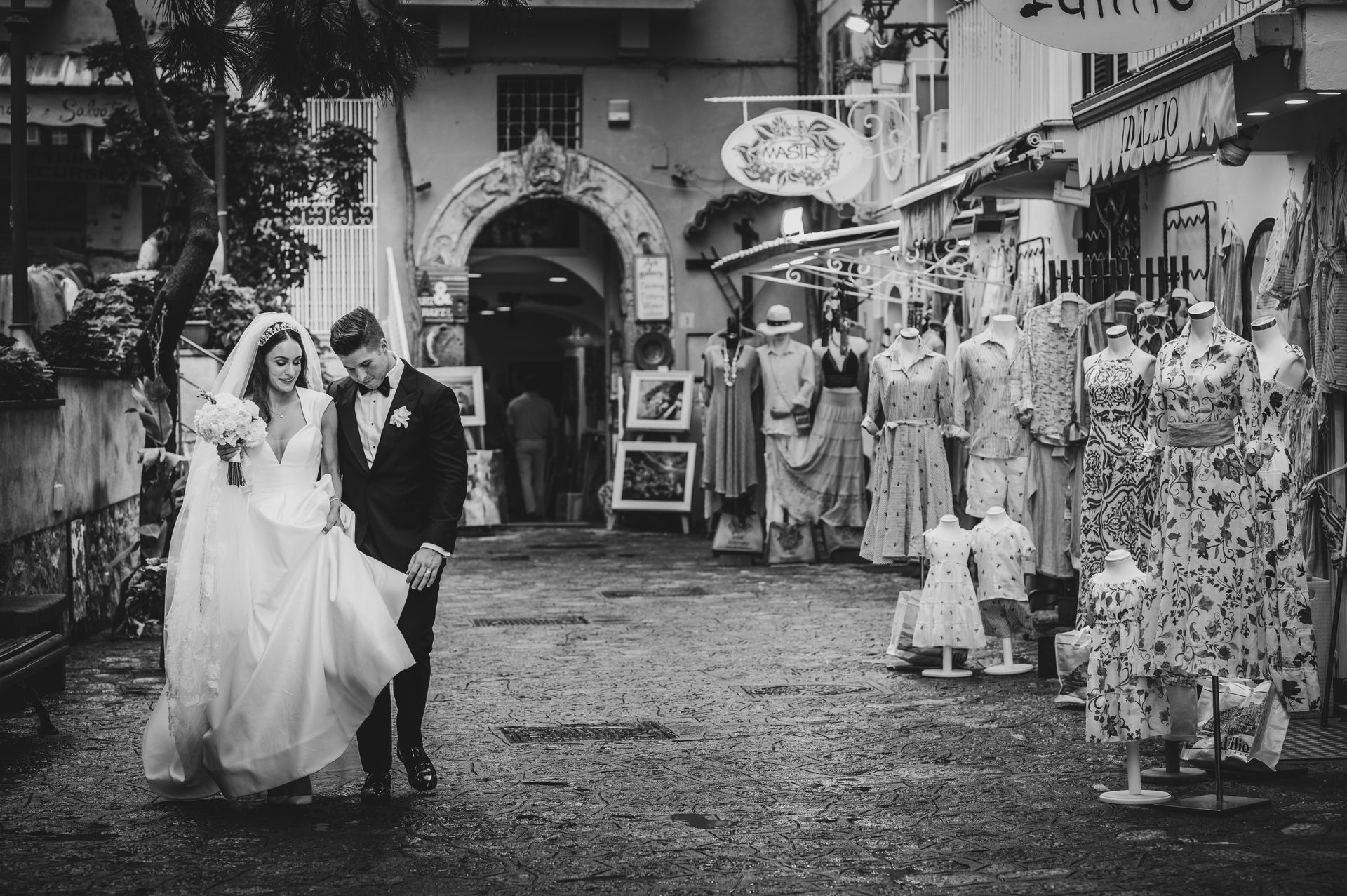 A bride and groom walk hand-in-hand along a narrow, stone-paved European street lined with boutique shops.