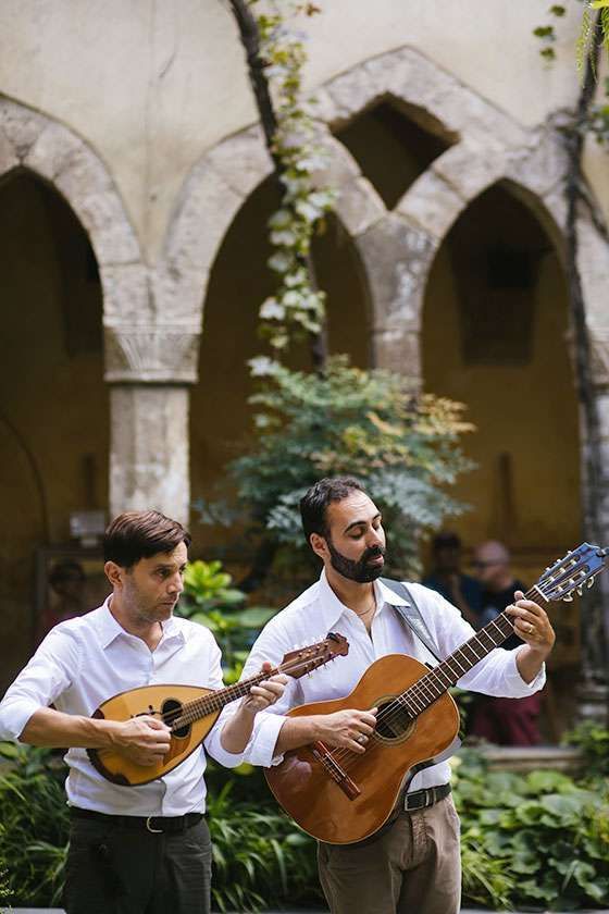 Two musicians in white shirts playing a mandolin and an acoustic guitar in a stone-arched garden courtyard.