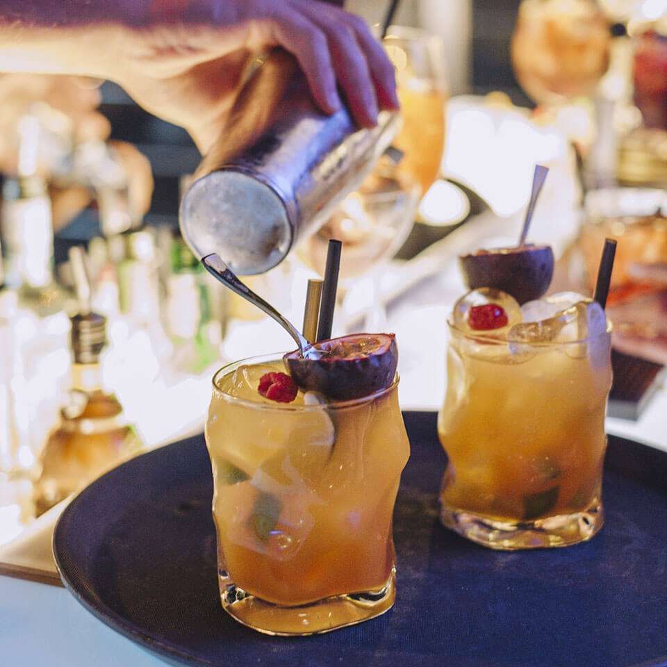 Un barista versa un cocktail da uno shaker in un bicchiere guarnito con mezzo frutto della passione e un lampone, posto su un vassoio.