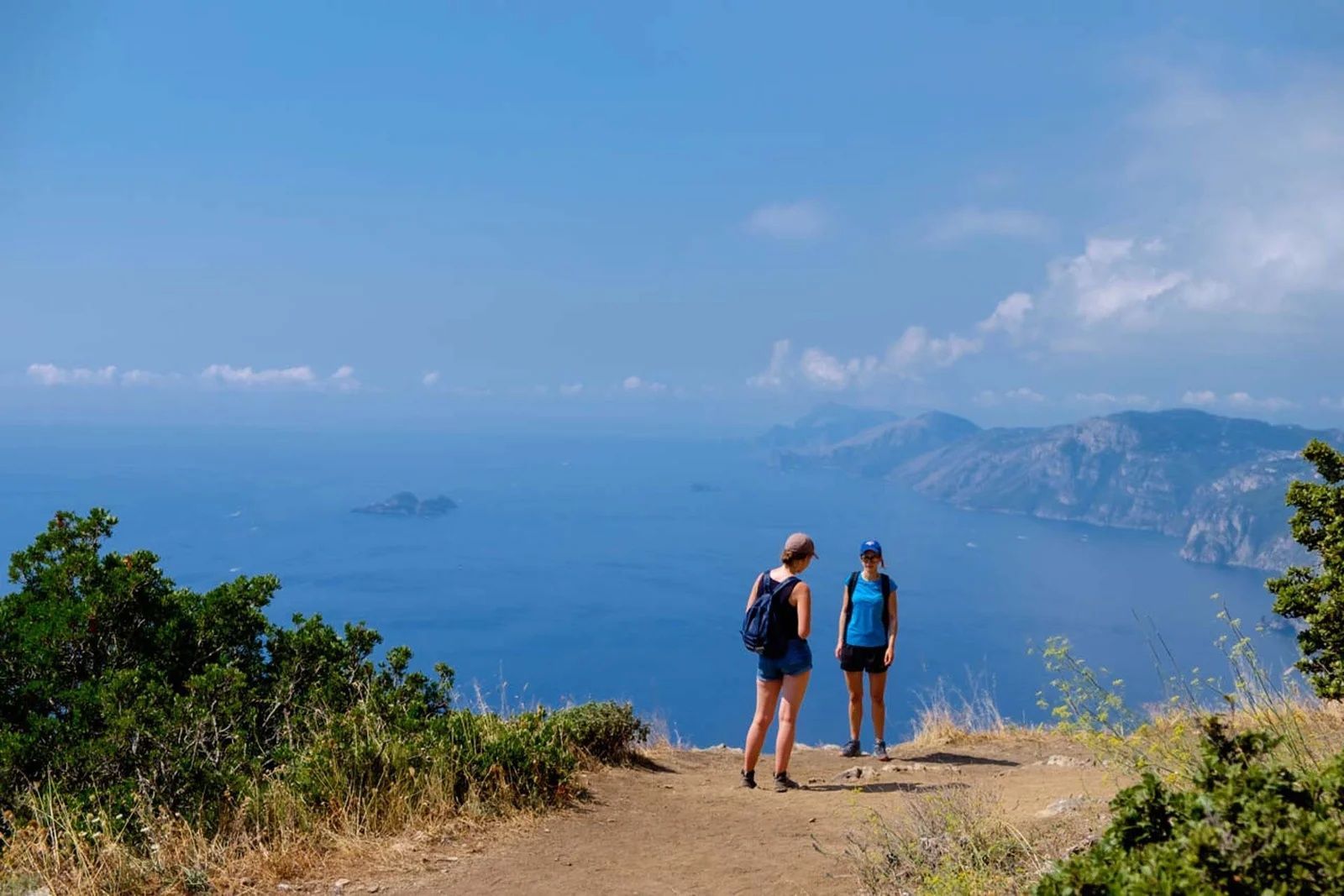 Two hikers stand on a sunlit coastal cliff overlooking the deep blue sea and distant mountainous coastline.