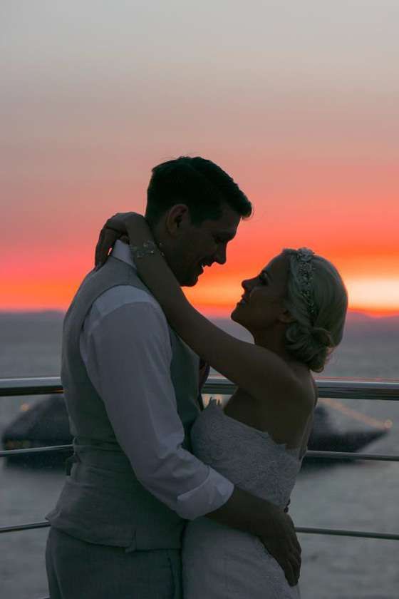 A couple in wedding attire embracing on a deck during a vibrant orange sunset over the ocean.