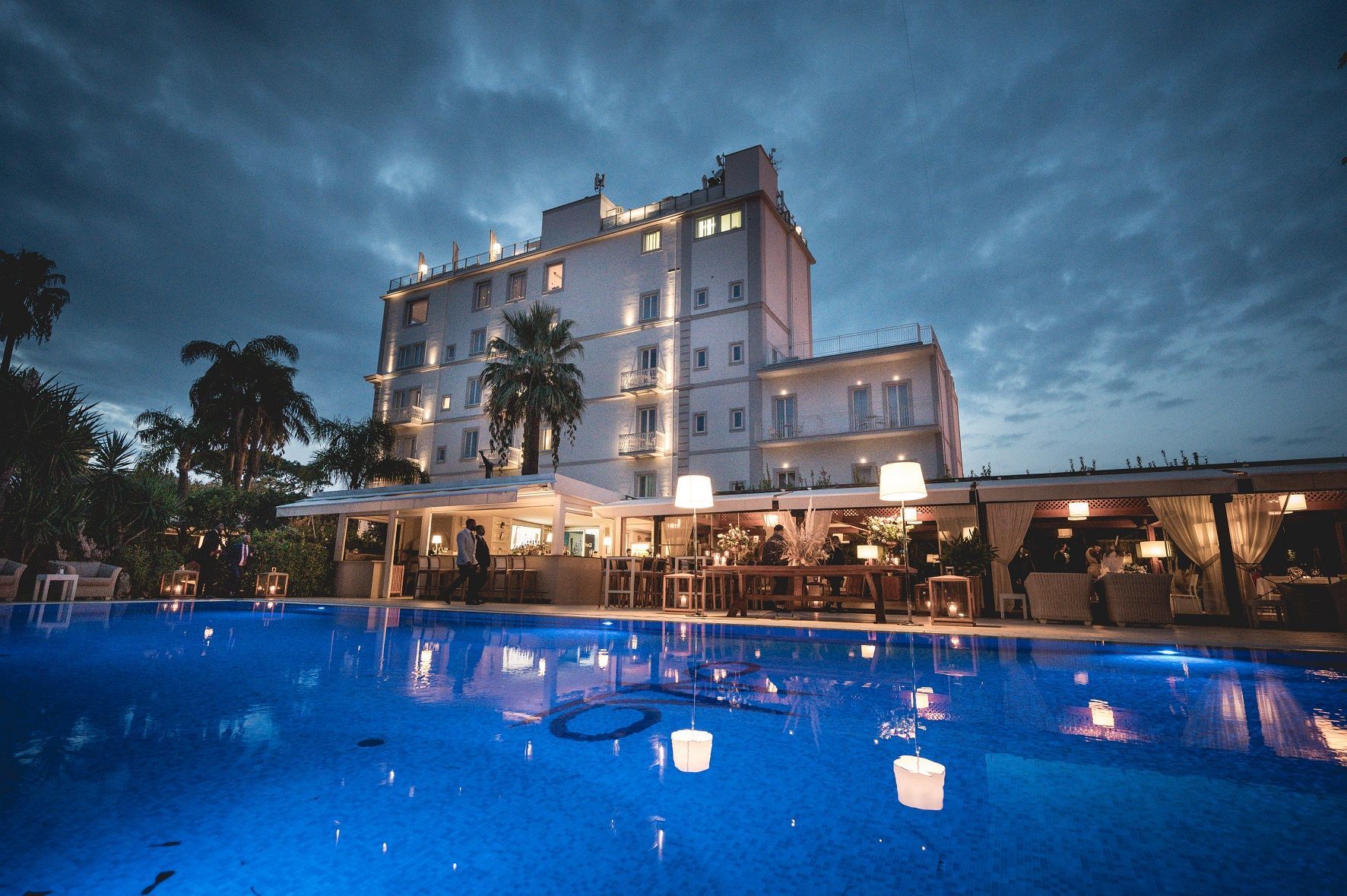 A tall, illuminated hotel at twilight, featuring a glowing blue swimming pool in the foreground and outdoor seating.