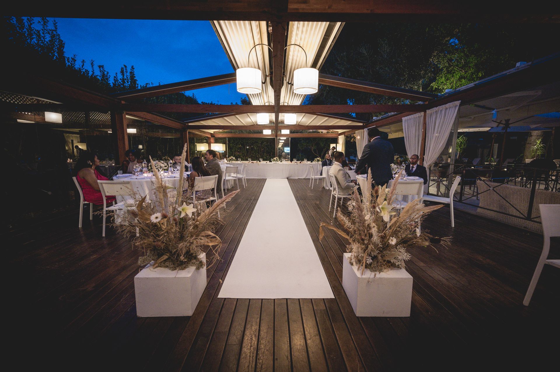 A white aisle runner leads to a long dining table under a wooden pergola at a twilight outdoor event.