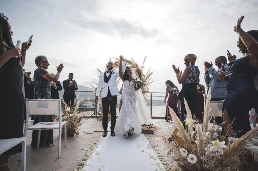 A bride and groom walk down a ceremony aisle together, hands raised in celebration as guests clap and cheer outdoors.