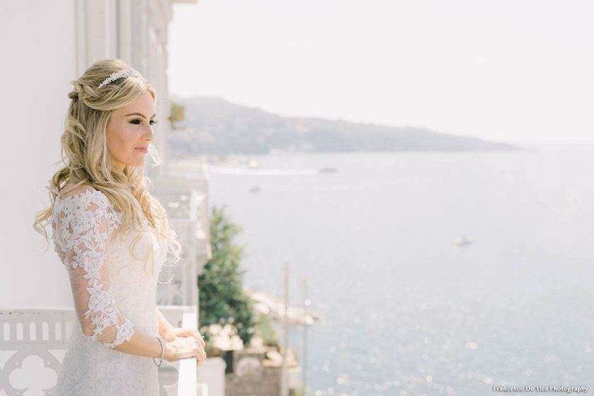 A blonde bride in a lace-sleeved gown stands on a balcony overlooking a bright, hazy coastline.