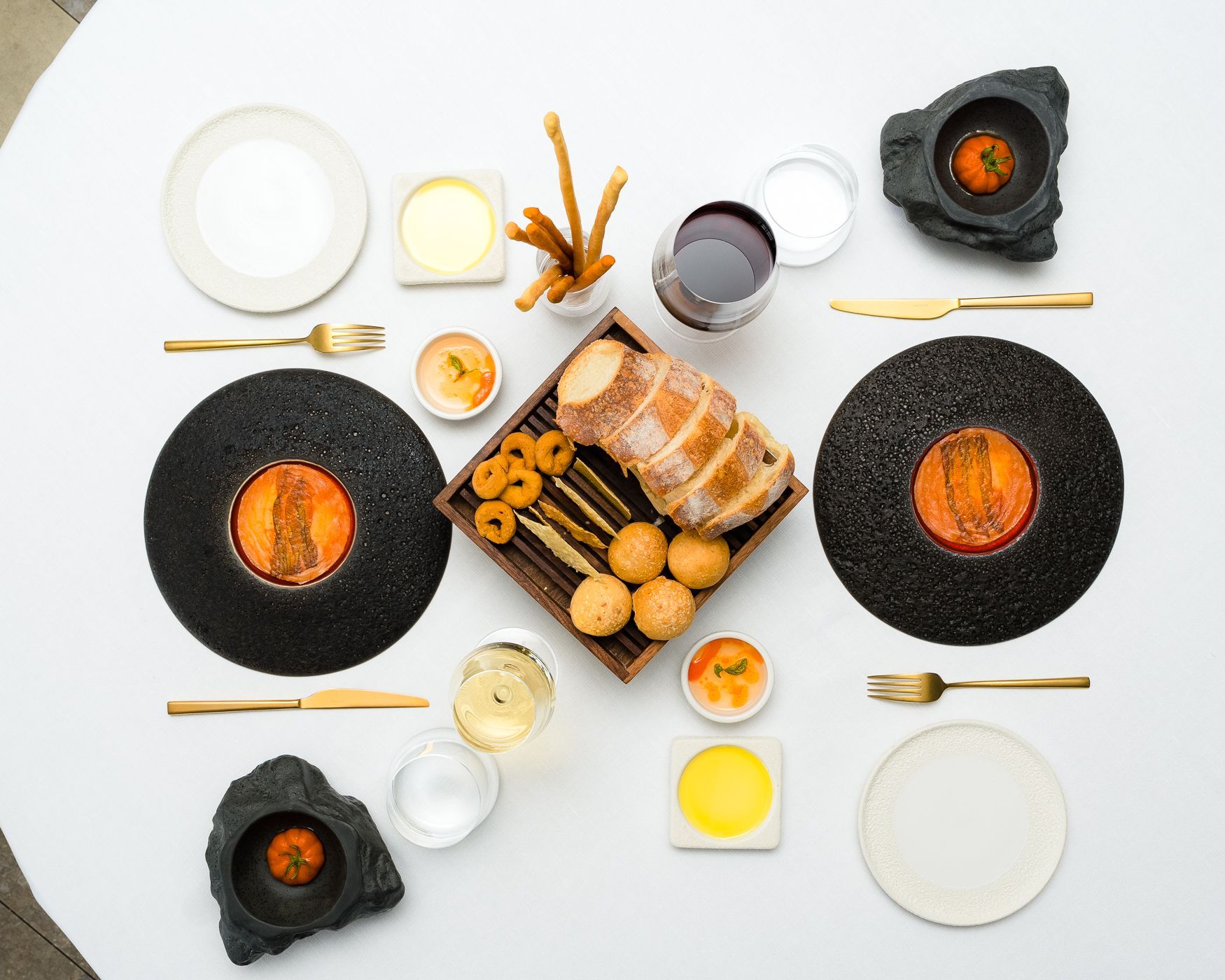 A gourmet meal spread on a white table featuring two black textured plates, bread, appetizers, and glasses of wine.