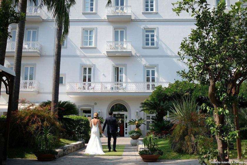 A bride and groom walk hand-in-hand toward a white, multi-story hotel entrance surrounded by lush gardens.