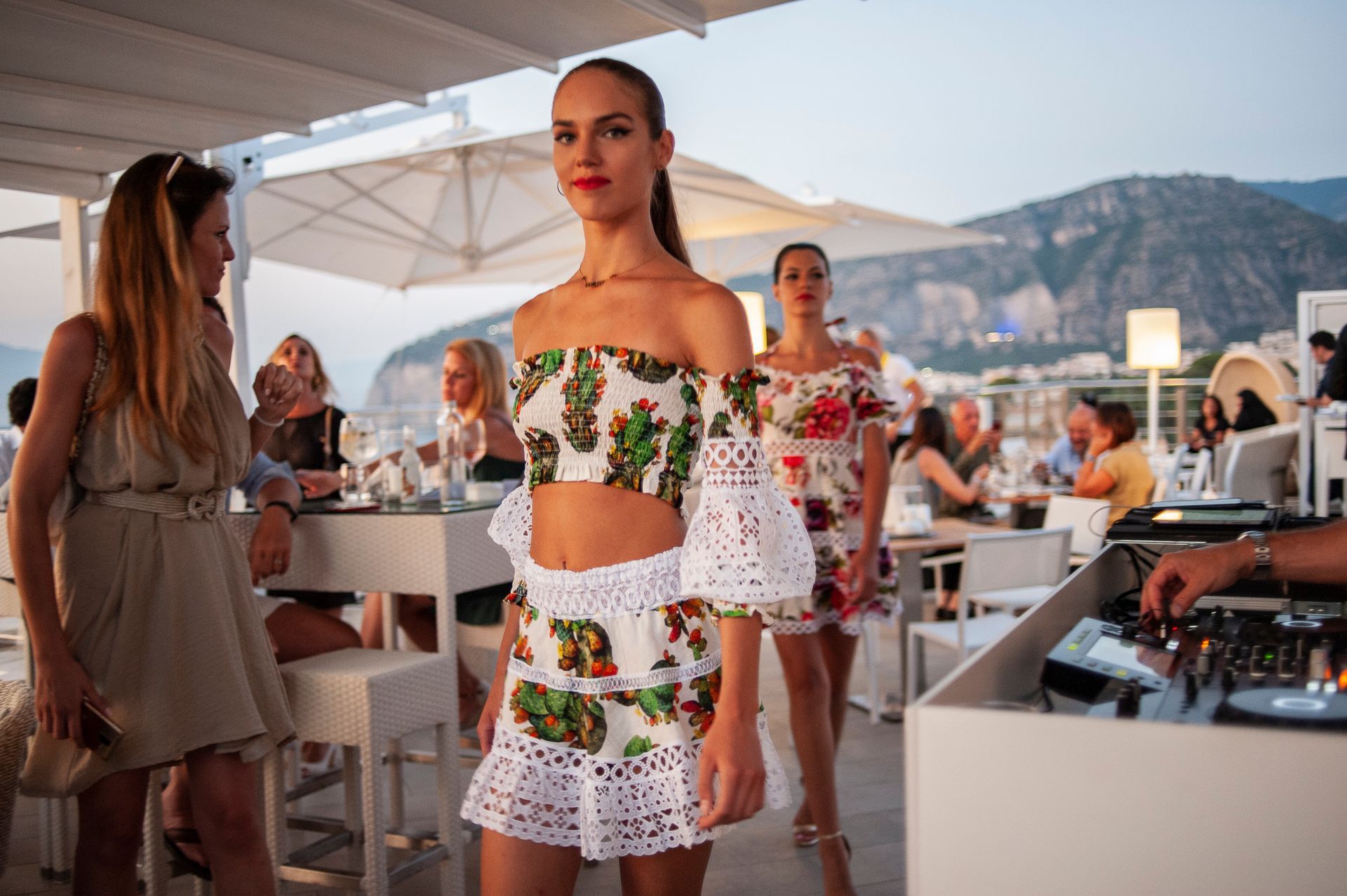 Models walk on a runway at an outdoor rooftop bar featuring tables, white umbrellas, and a sunset mountain backdrop.