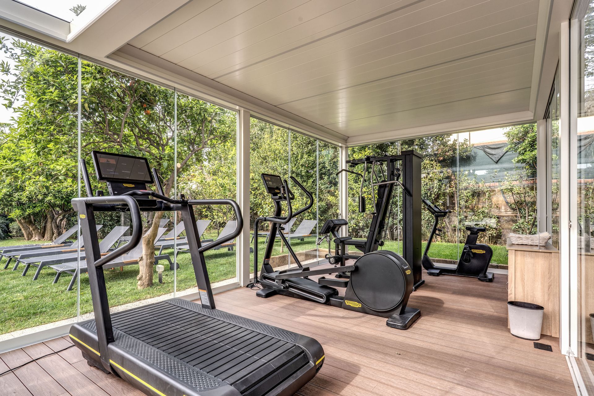 Una moderna palestra domestica in una stanza vetrata chiusa, dotata di tapis roulant, ellittica e cyclette su una piattaforma di legno.