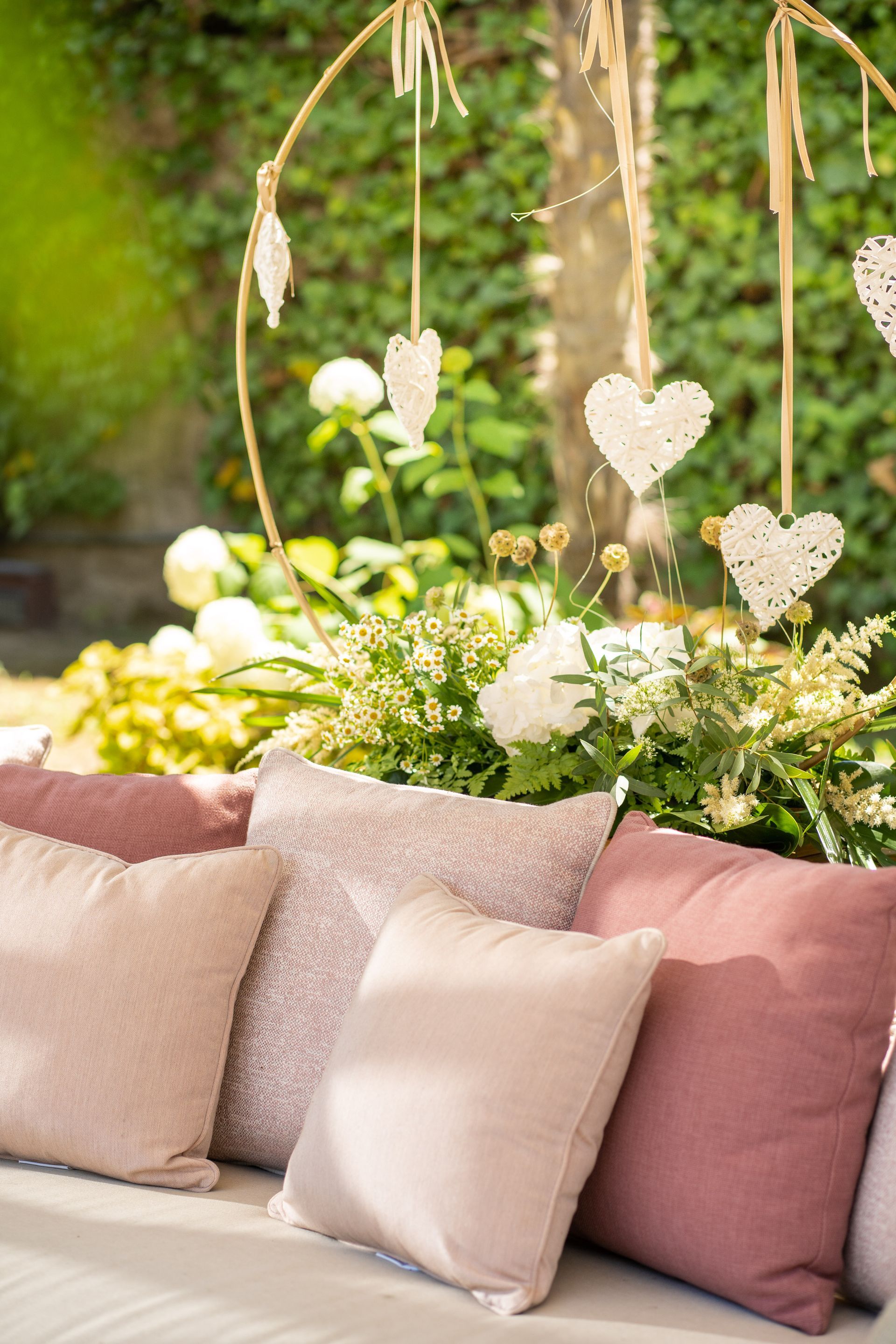 A row of soft pink and textured cushions sits before a garden display of white flowers and hanging decorative hearts.