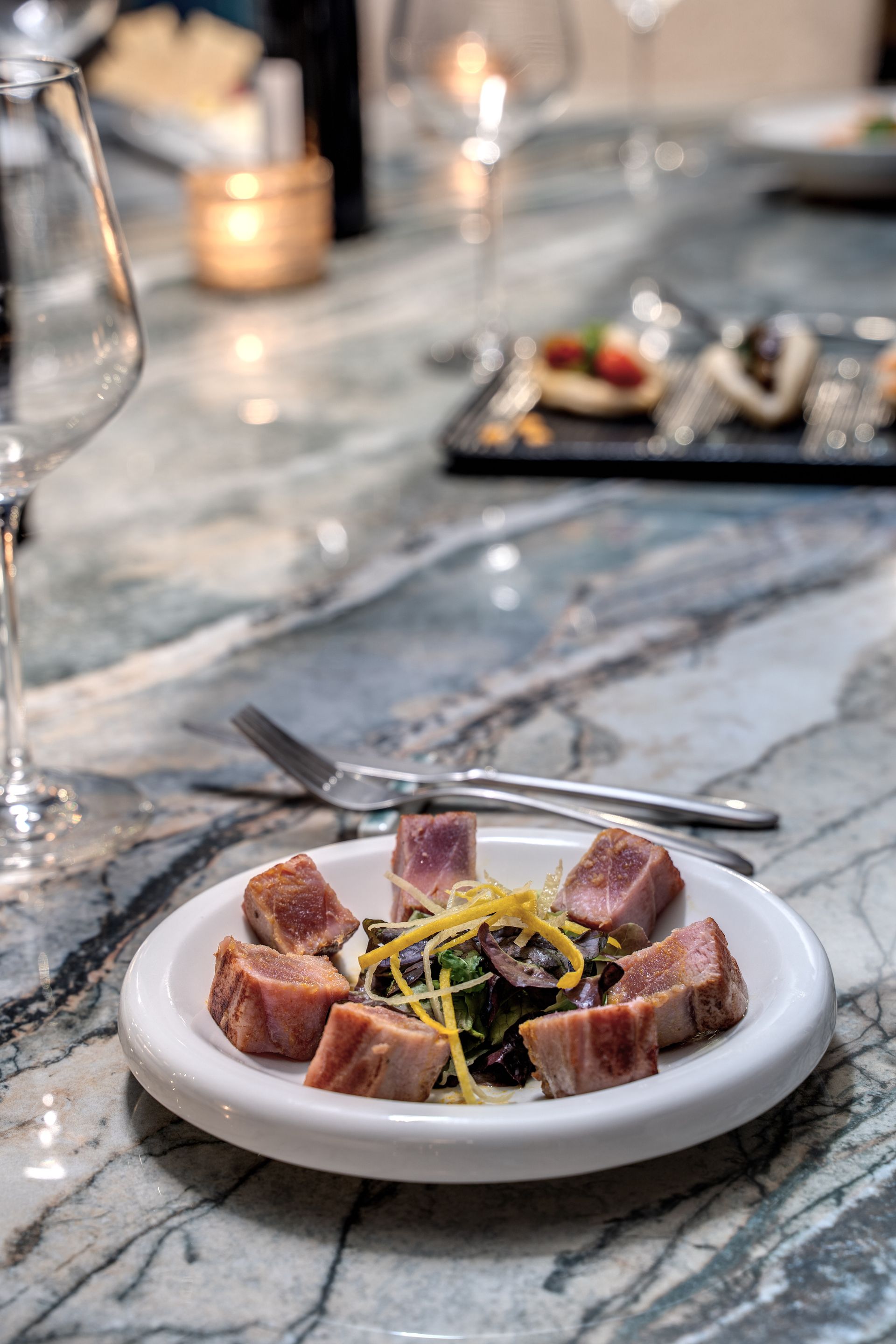 Su un tavolo di marmo grigio, accanto a un calice di vino e a una tavola apparecchiata in modo sfocato, si trova un piatto bianco con cubetti di tonno scottato e verdure.