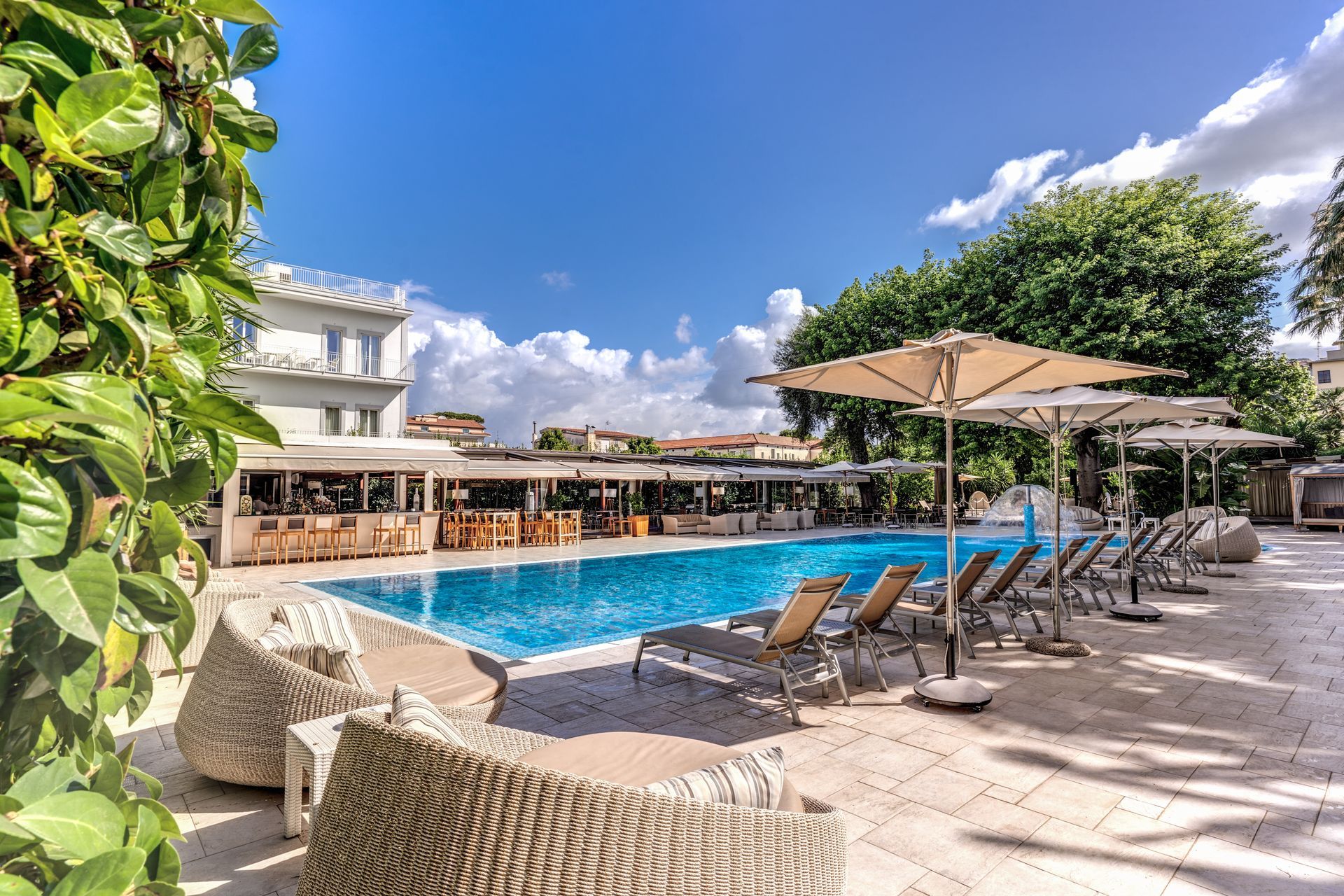 A resort swimming pool surrounded by lounge chairs, patio umbrellas, and lush greenery under a bright, sunny sky.