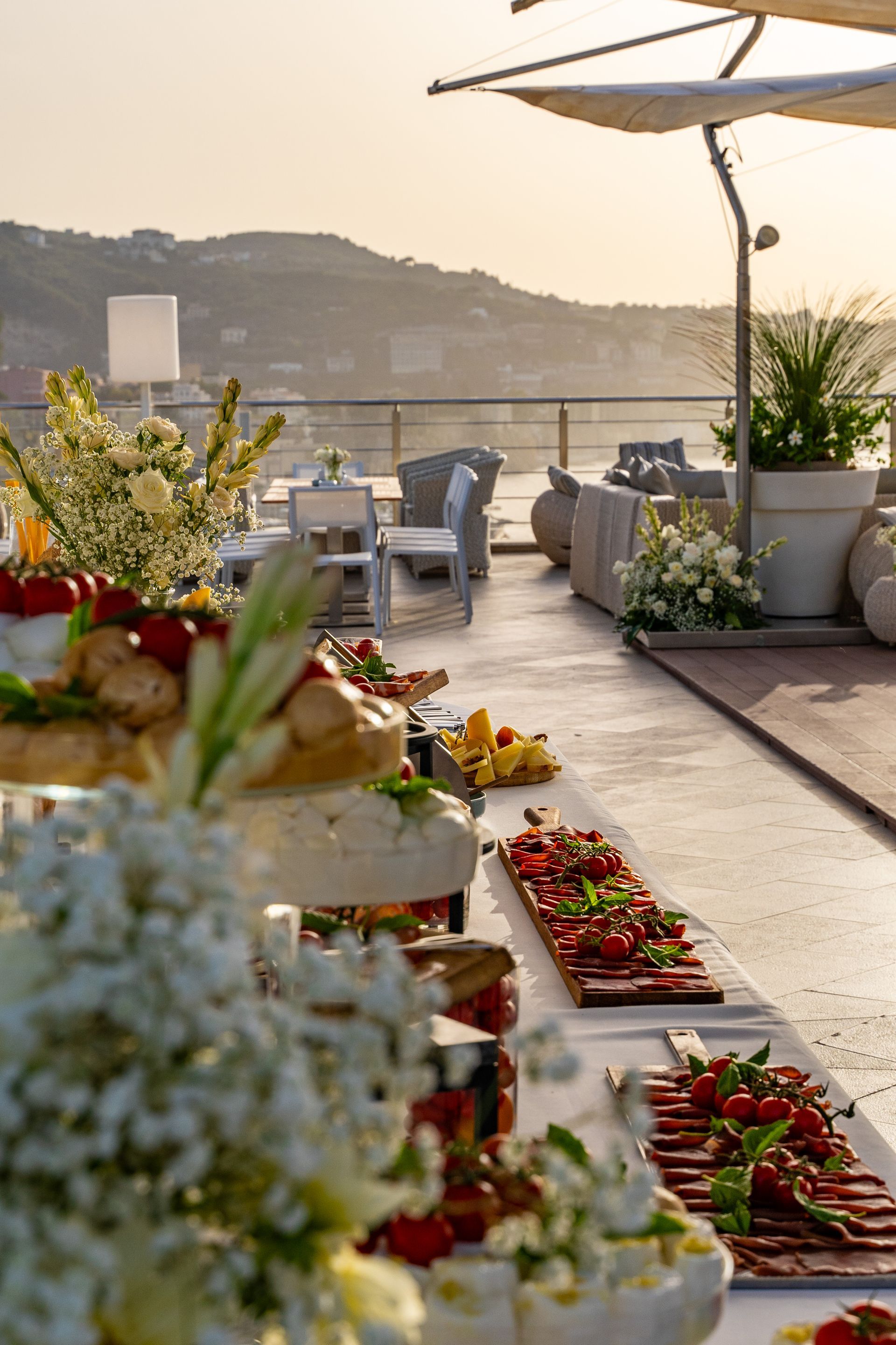 A sunset terrace setup with a buffet of appetizers arranged on tables, overlooking a sea view.