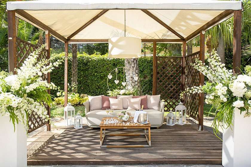 A wooden pergola covers a lounge area with a sofa, coffee table, hanging lamp, and floral arrangements in a lush garden.