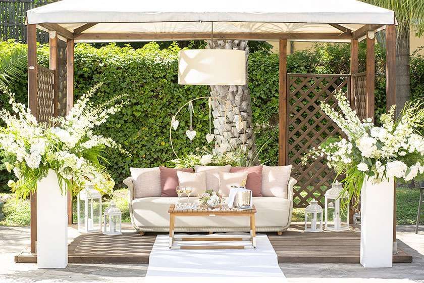 An outdoor wedding lounge with a beige sofa, wooden coffee table, and large white floral arrangements under a canopy.