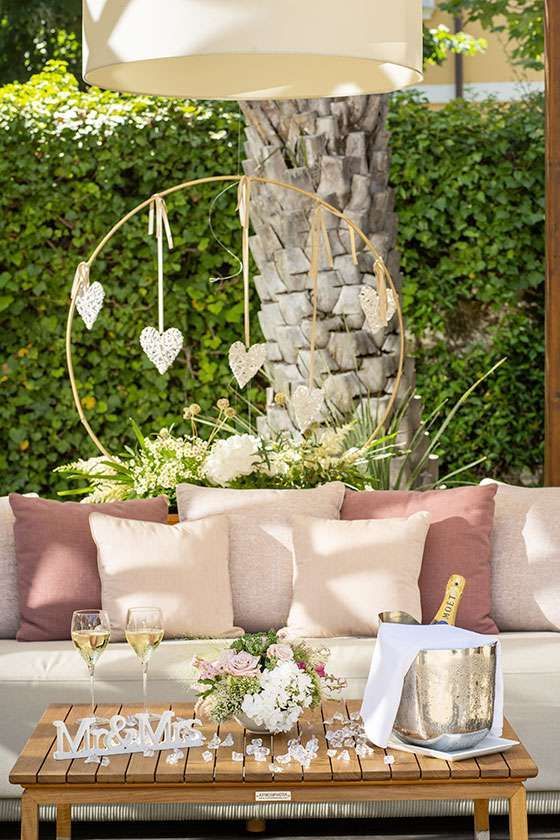 A lounge setting featuring a sofa with pink cushions, a wooden table with champagne, and a gold hoop with hanging hearts.