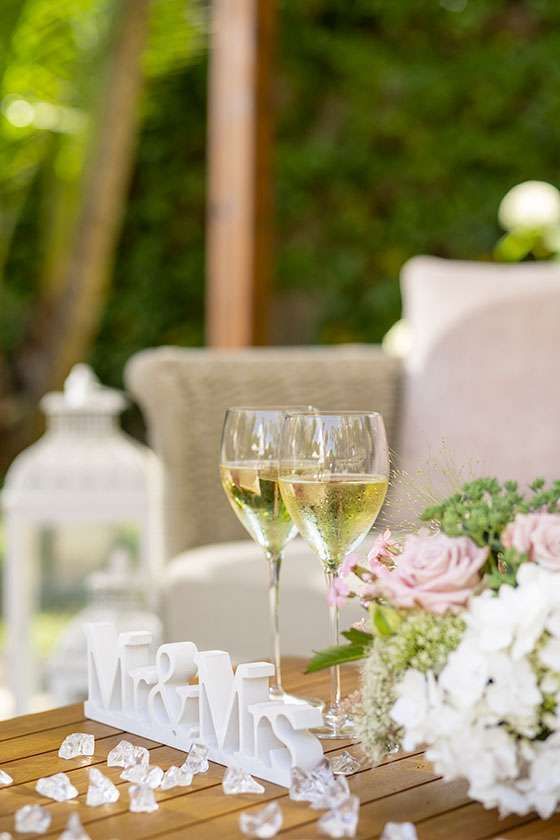 Two glasses of champagne sit on a wooden table beside a floral arrangement and a white 
