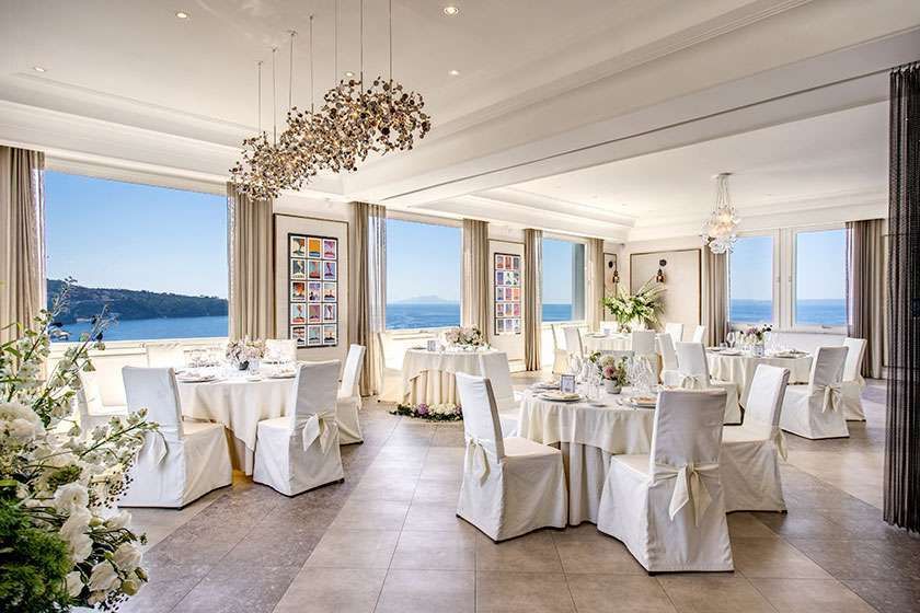 Bright dining room with white-clothed tables, chairs with tied bows, and large windows overlooking the blue ocean.