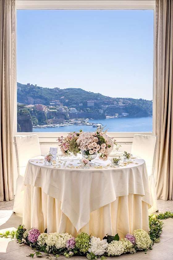 A small, cream-colored dining table decorated with floral arrangements, set before a window overlooking a coastal town.