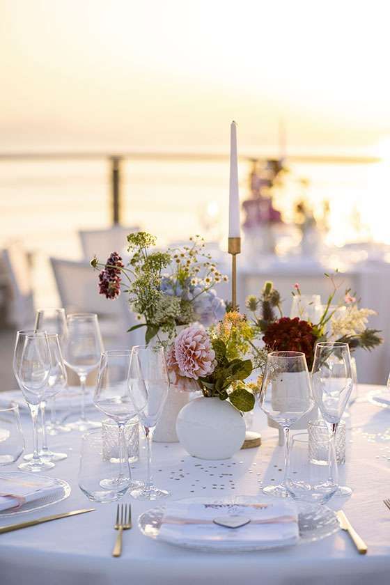 A floral centerpiece with a tall candle sits on an elegant table set for dinner at sunset, overlooking the ocean.
