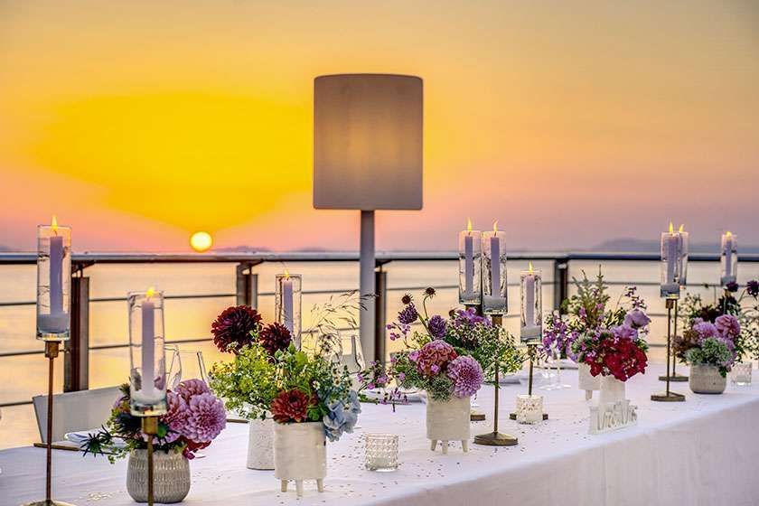 A long table set with floral arrangements and tall pillar candles at sunset overlooking a calm ocean.