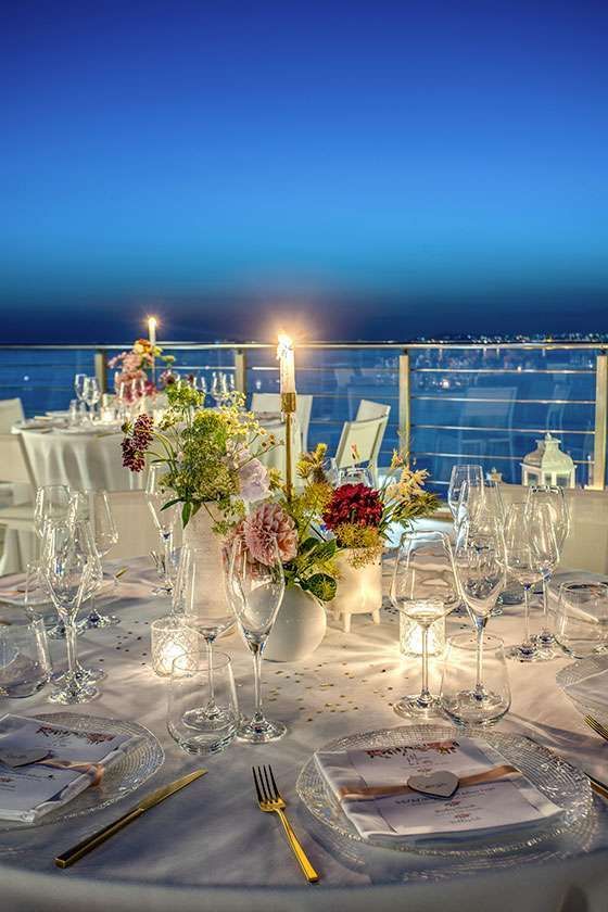 A beautifully set dining table with flowers and candles at dusk, overlooking a vast, dark sea under a clear blue sky.
