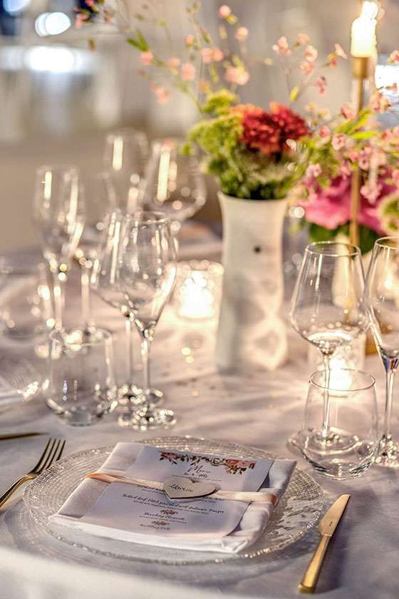 Elegant table setting with a white vase of flowers, crystal glasses, and a place card on a charger plate.