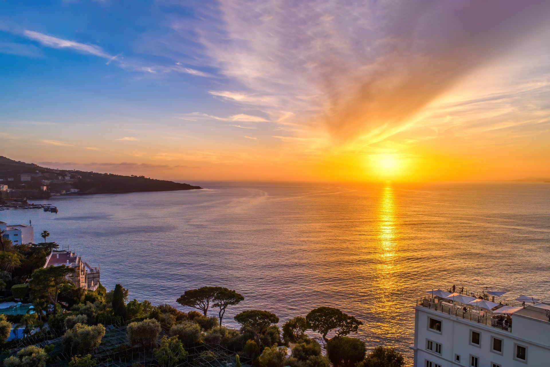 A vibrant sunset glows over a calm ocean, with a coastal hillside and a white building nestled among trees in the foreground.