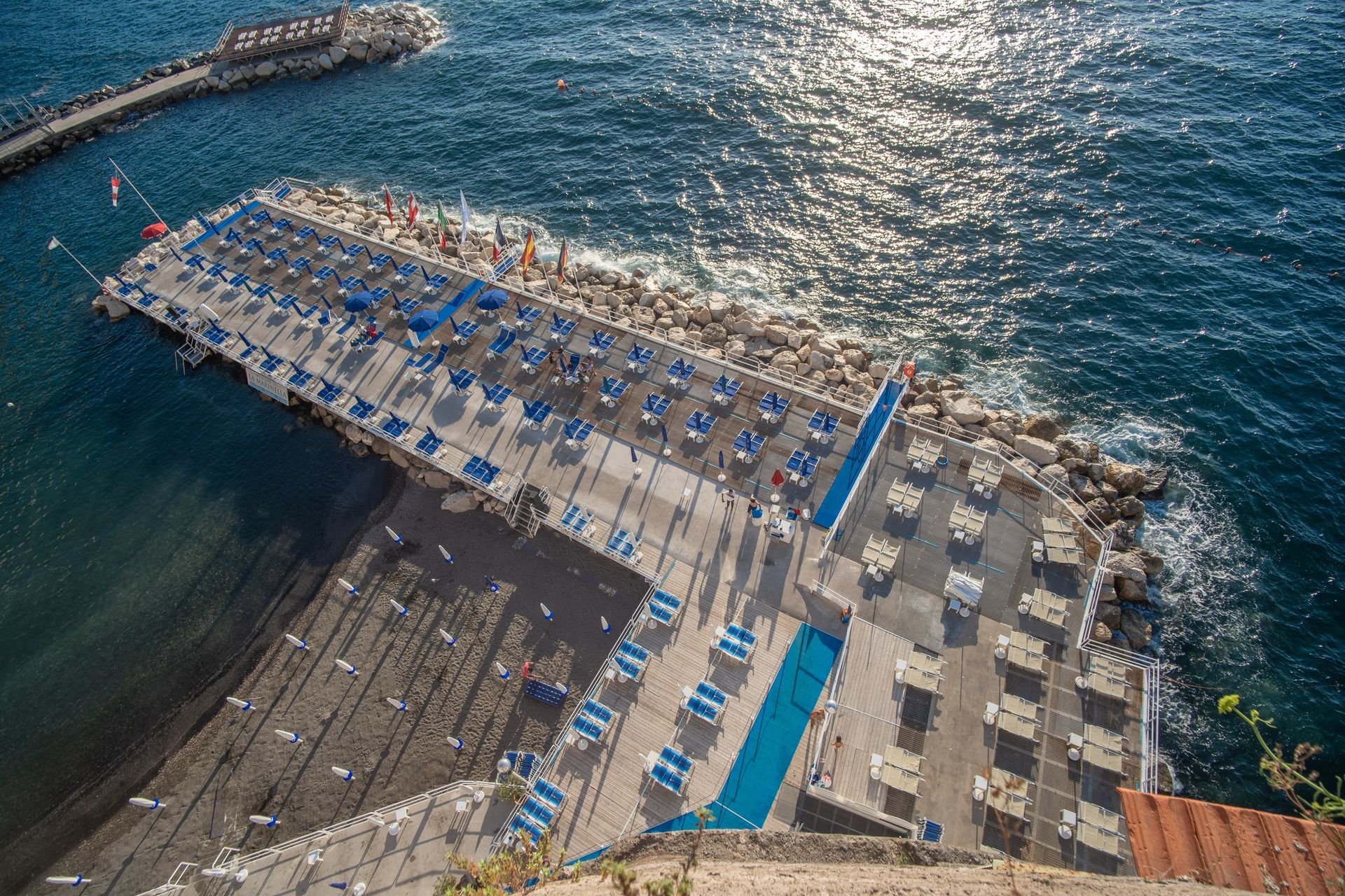 Veduta aerea di un beach club costiero con file di lettini blu e bianchi su un molo pavimentato circondato dal mare.