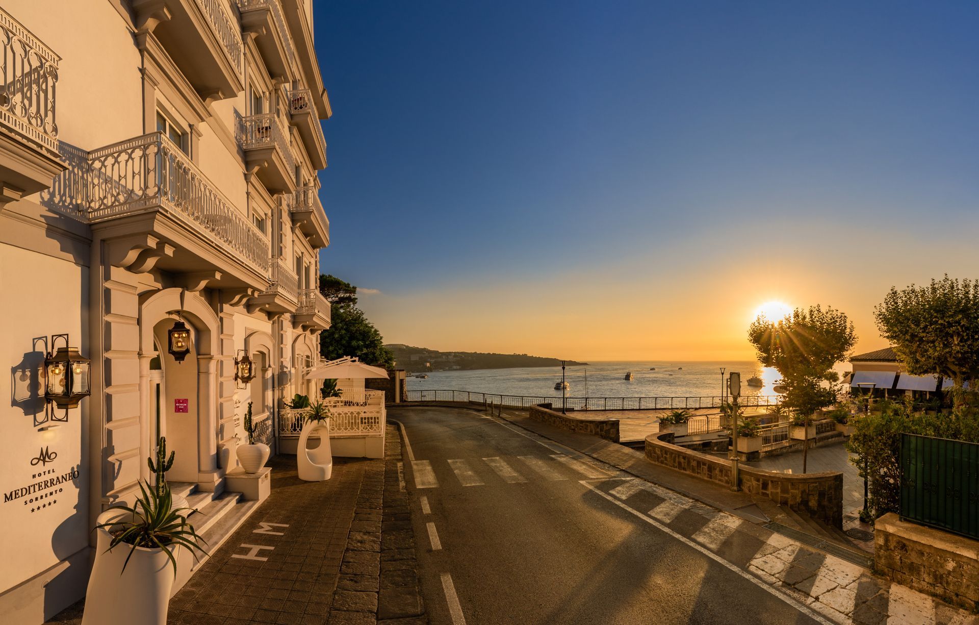 Sunset over the sea beside a grand, multi-story hotel with decorative balconies and a coastal road.