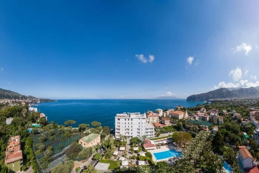 Aerial view of a white hotel resort by a deep blue sea under a clear sky, with surrounding green trees and buildings.