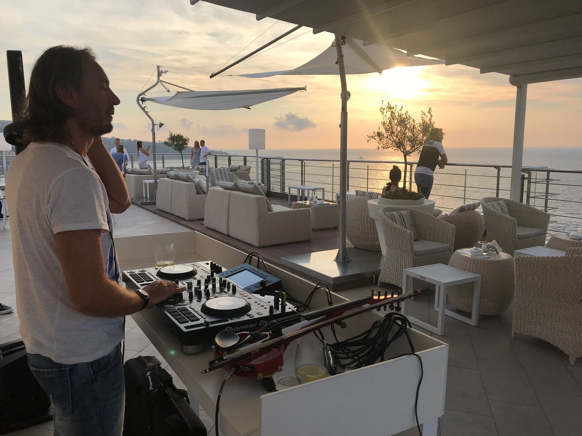 A DJ at a mixing console on an outdoor seaside terrace during sunset, with people seated at tables nearby.