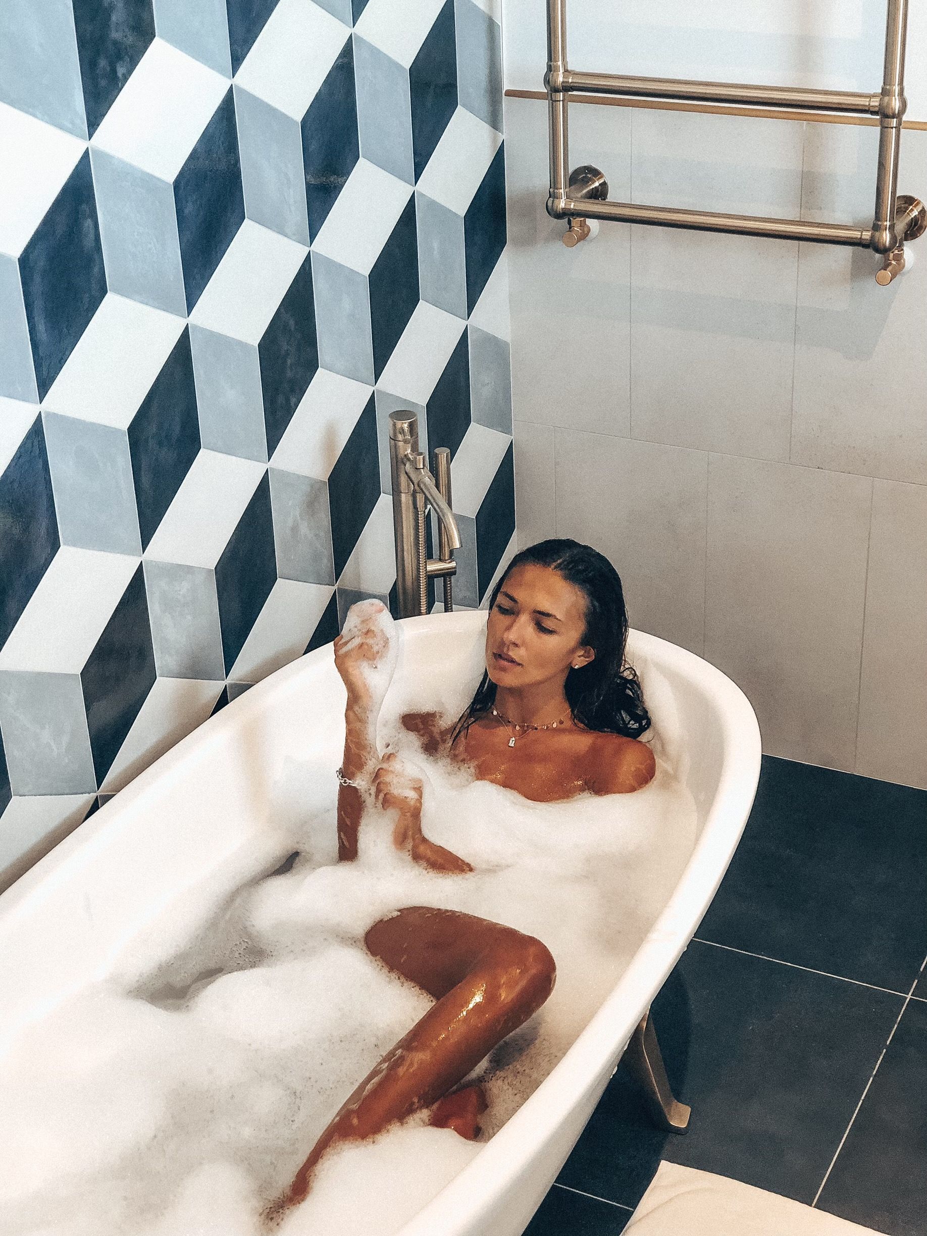 A person with dark hair sits in a white clawfoot bathtub filled with soap bubbles, against a patterned geometric wall.
