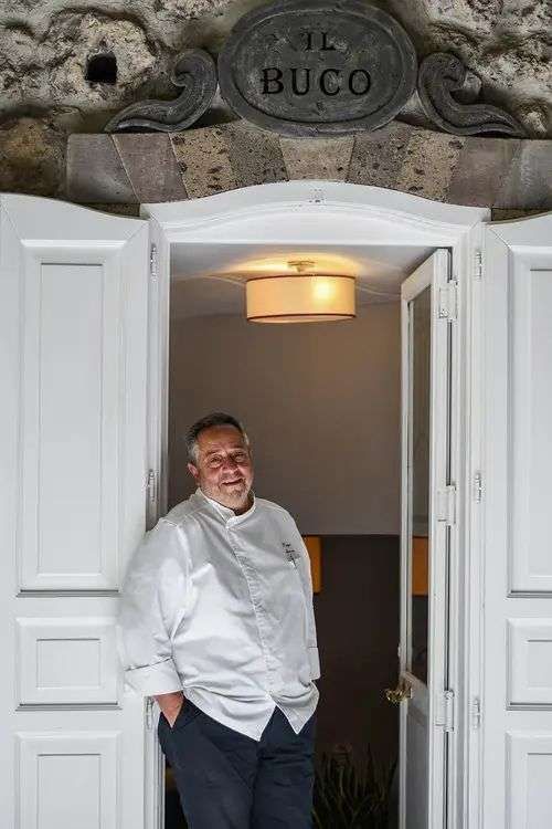 Uno chef in uniforme bianca sorride sulla soglia di un ristorante con l'insegna 