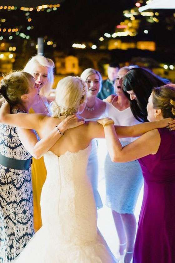 A bride in a white wedding dress stands in a circle with friends who have their arms around each other at night.