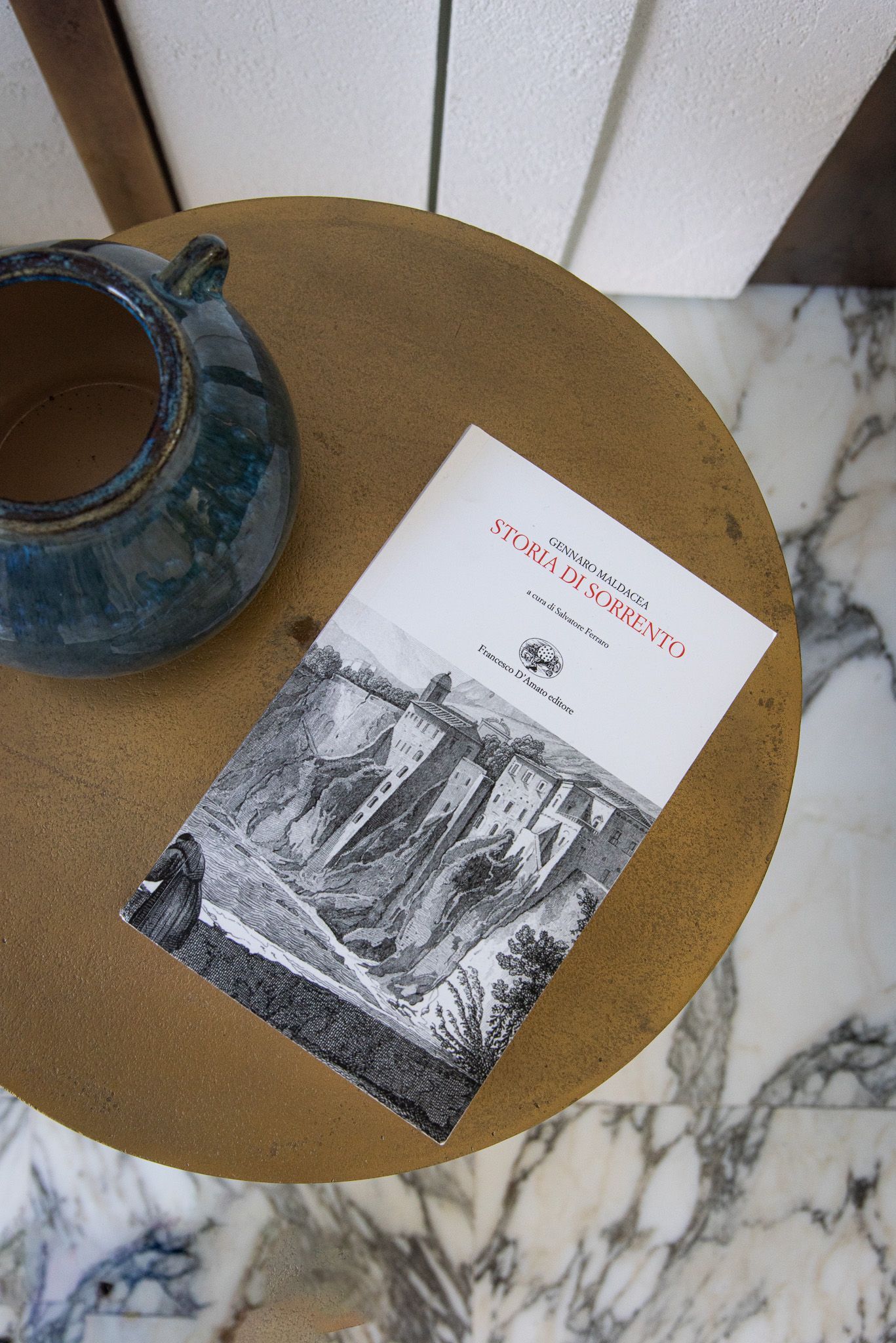 Un piccolo libro con una copertina raffigurante un paesaggio in bianco e nero è appoggiato su un tavolo rotondo di legno accanto a una brocca di ceramica blu.