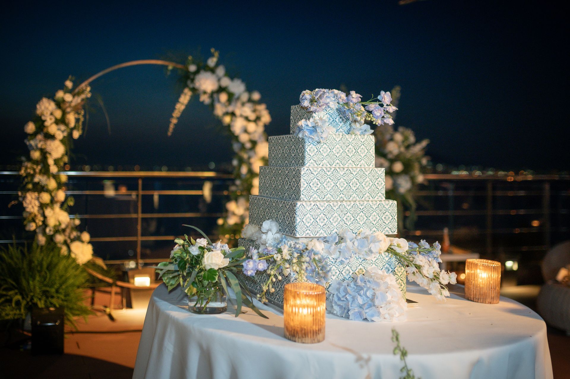 A four-tier, patterned wedding cake decorated with blue flowers, displayed on a table outdoors at night with an arch.