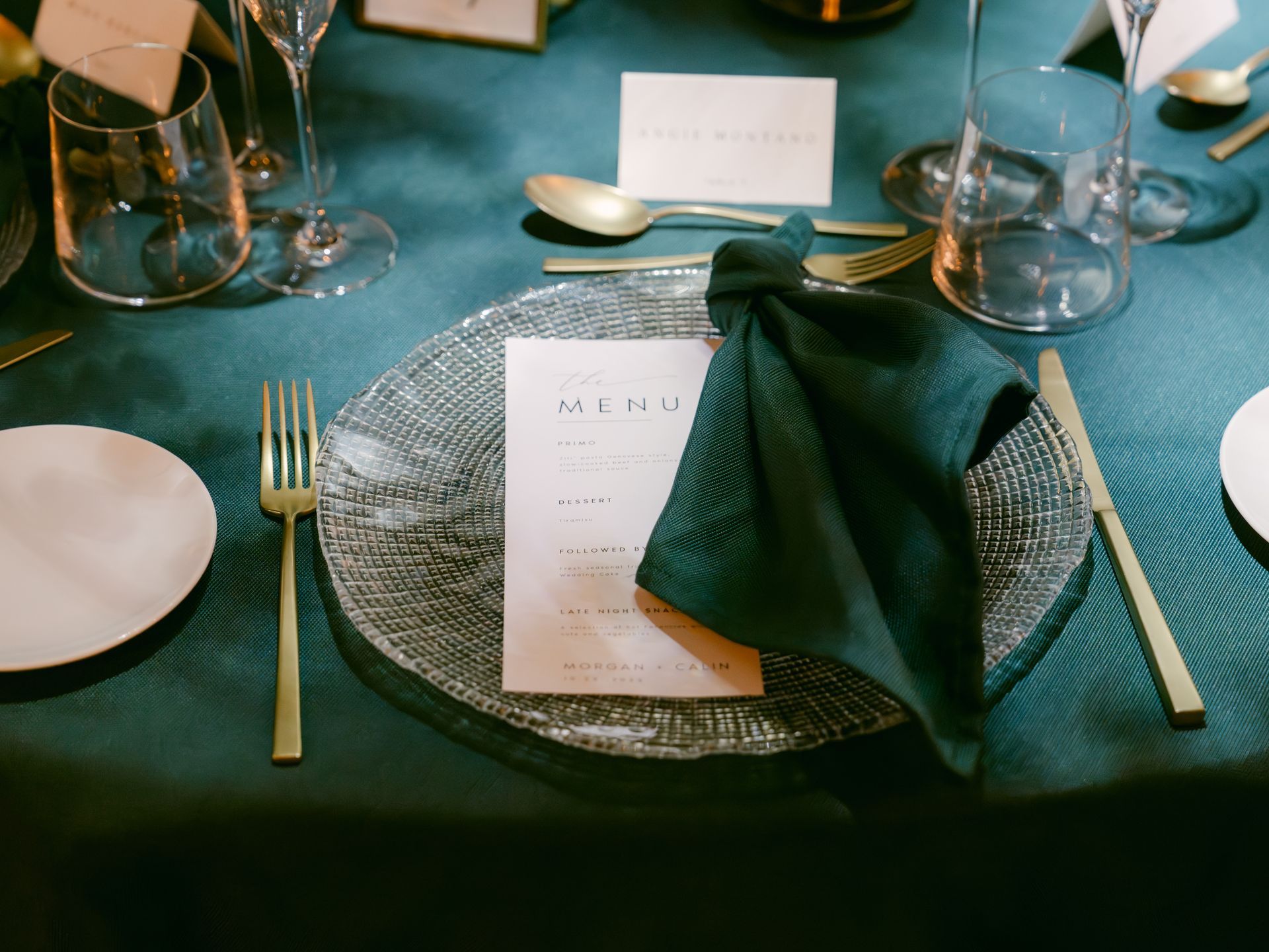 A formal place setting with a dark green napkin, menu card, and gold silverware on a teal velvet tablecloth.