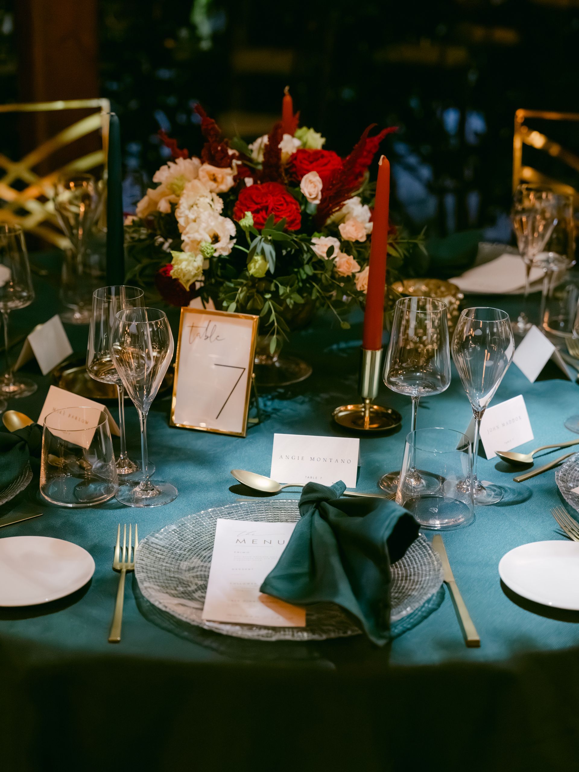 A festive wedding table setting with a dark teal tablecloth, red floral centerpiece, taper candles, and place cards.