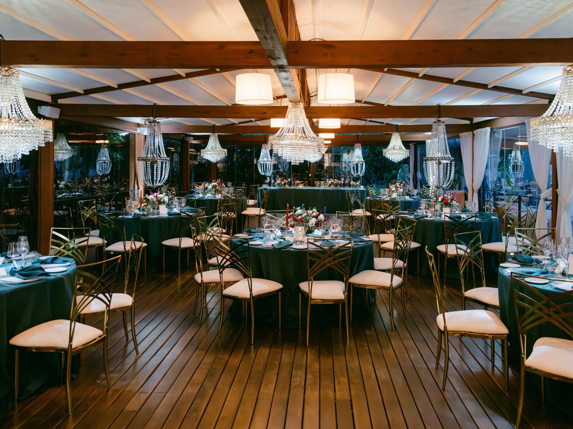 Elegant banquet room with round tables, dark green tablecloths, gold chairs, and crystal chandeliers under wood beams.