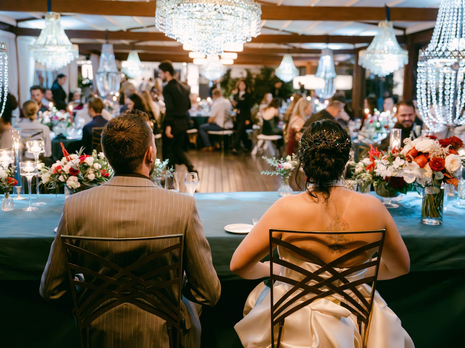 A newlywed couple sits at a head table at a wedding reception, viewed from behind in a candlelit, chandelier-lit venue.