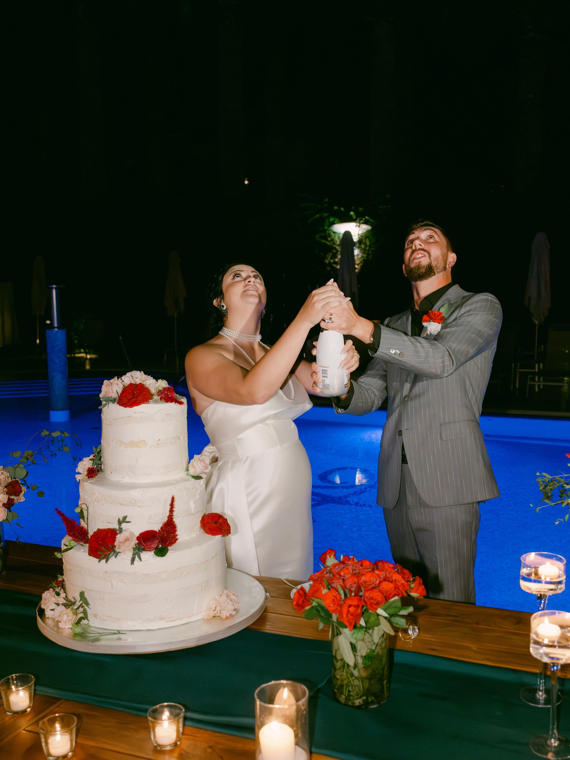 A bride and groom cut a tiered white wedding cake adorned with red roses by a pool illuminated with blue light at night.