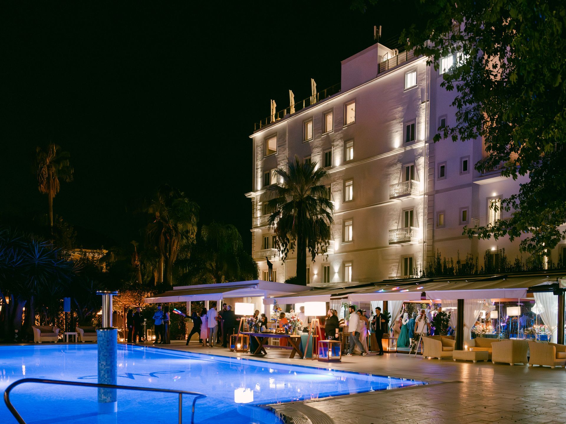 A night scene of a lit hotel exterior by a glowing blue swimming pool, with people gathering on the terrace.