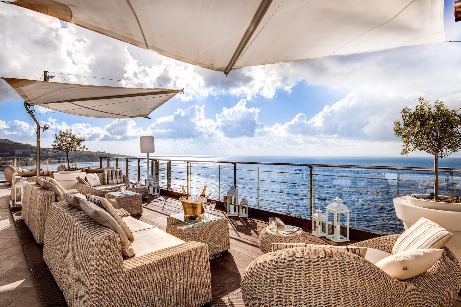 A luxury seaside terrace featuring wicker sofas, white umbrellas, and decorative lanterns under a bright, cloudy sky.