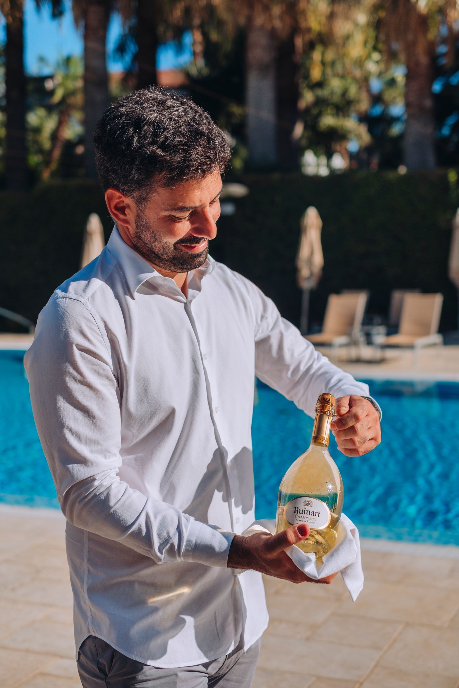In una giornata di sole, una persona con una camicia bianca abbottonata stappa con cura una bottiglia di vino a bordo piscina.