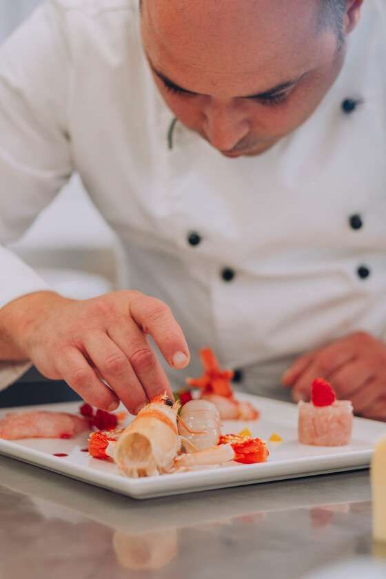 Uno chef in uniforme bianca guarnisce con cura un piatto di pesce pregiato, impiattato con eleganza su un piatto quadrato bianco.